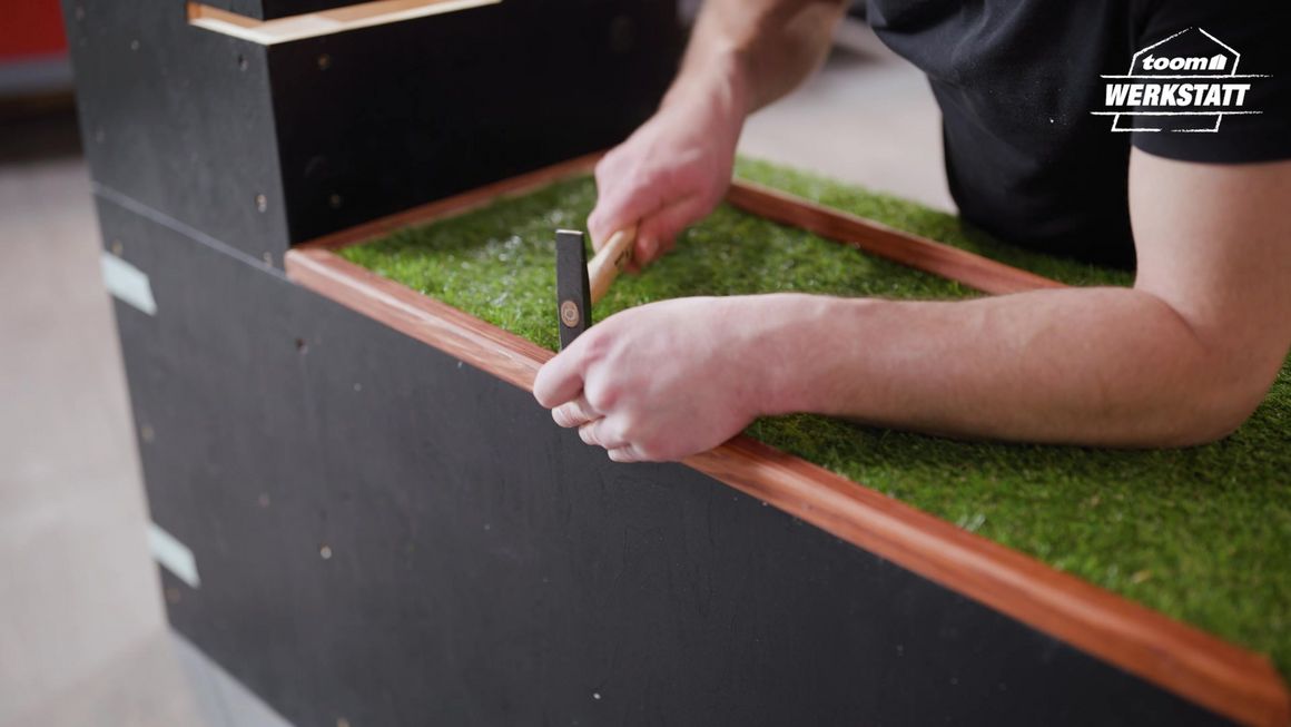 Person hämmert Holzleiste an Kiste mit Kunstrasen