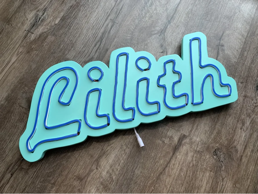 Ein blaues Neonschild mit dem Namen Lilith auf einem hellgrünen Hintergrund.