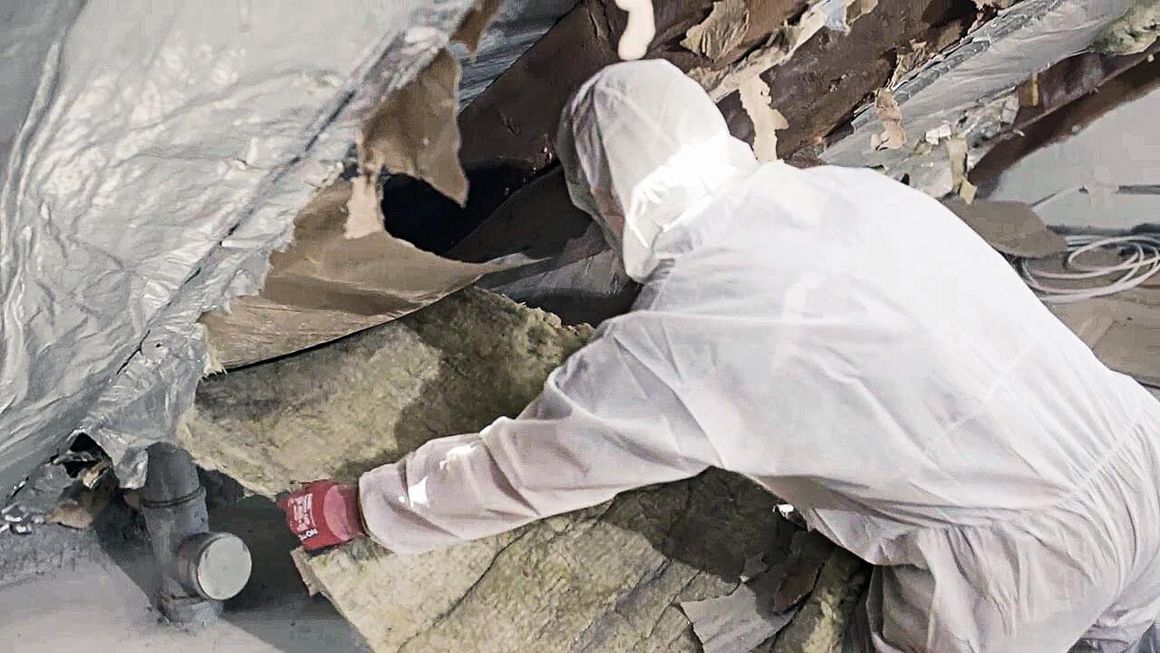 Handwerker in Schutzkleidung entfernt altes Dämmmaterial aus dem Dachstuhl.