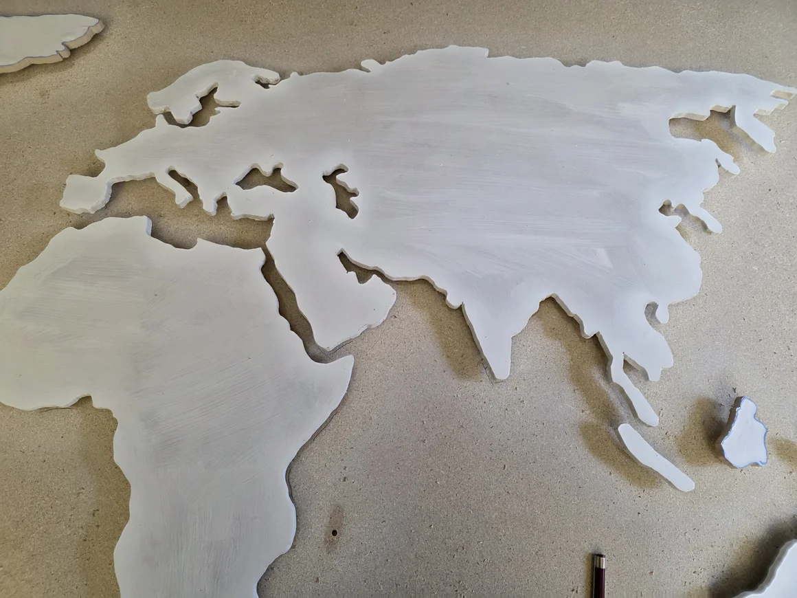 Eine weiß bemalte, ausgeschnittene Weltkarte liegt auf einer Holzplatte.