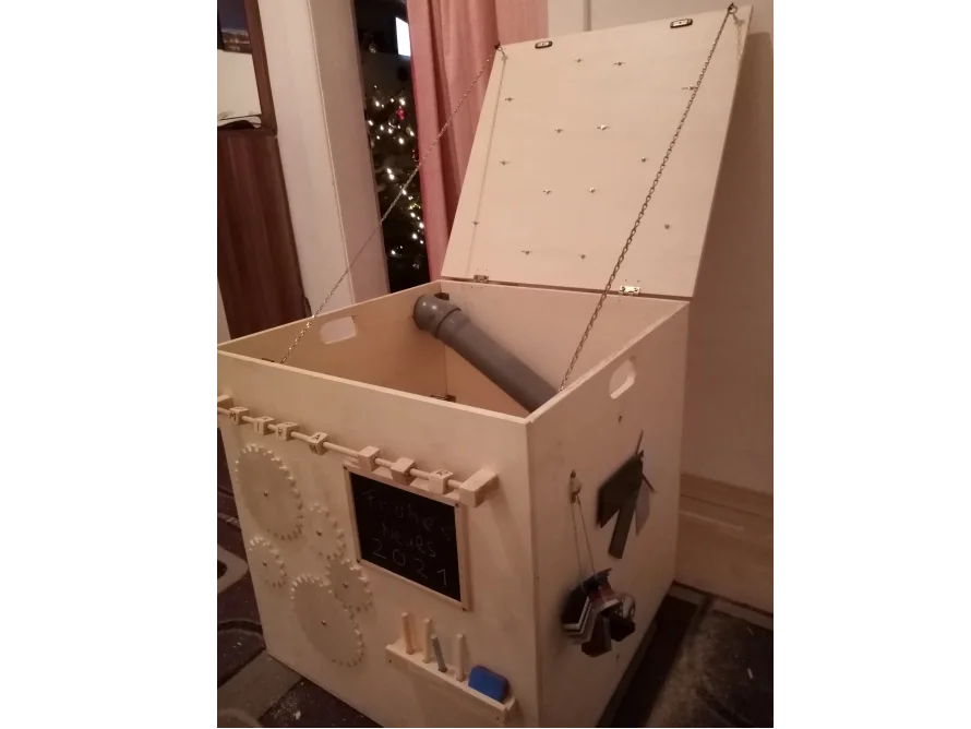 Eine Holzkiste mit Deckel, an der verschiedene motorische Spielelemente angebracht sind.