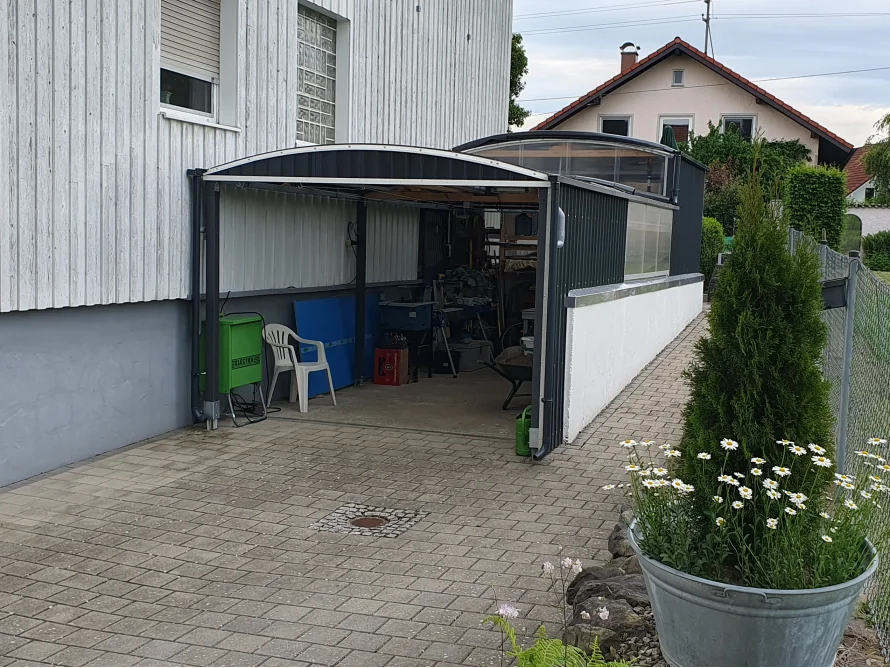 Ein angebauter Carport aus Metall neben einem gepflasterten Weg.