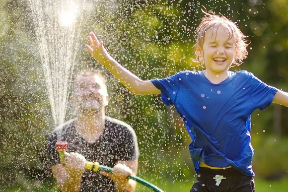 Person bespritzt fröhliches Kind mit Wasserschlauch im Garten