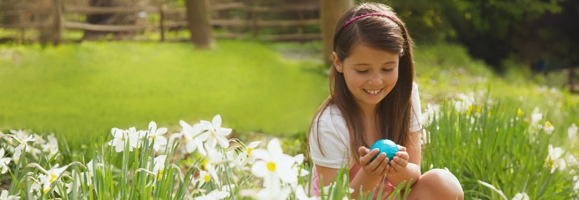 Ein lächelndes Kind hockt auf einer Wiese und hält ein blaues Osterei.