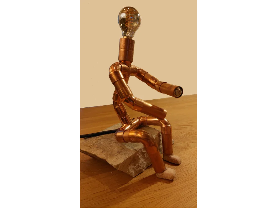 Eine aus Kupferrohren gefertigte Figur sitzt auf einem Stein.