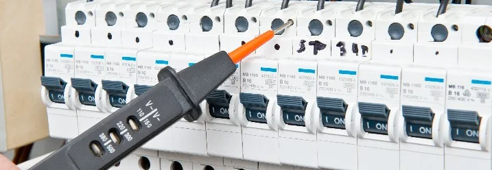 Nahaufnahme einer Hand, die einen Spannungsprüfer an einer Reihe von Sicherungen in einem Sicherungskasten hält.