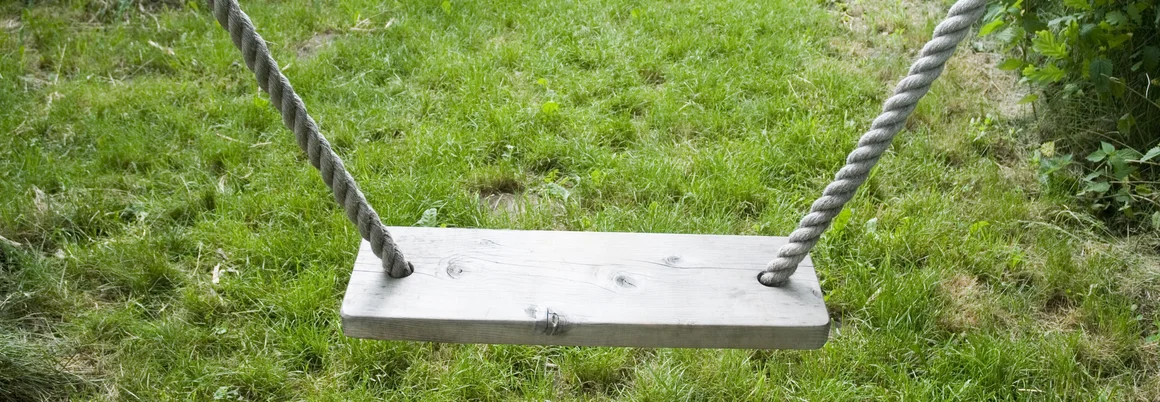 Eine leere Holzschaukel hängt an dicken Seilen über einer Wiese.