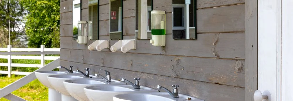 Mehrere weiße Waschbecken mit Spiegeln stehen an einer Holzwand im Freien.