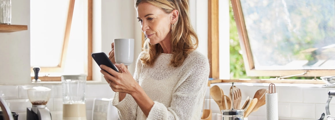 Eine Person schaut in der Küche auf ihr Smartphone und hält eine Tasse.