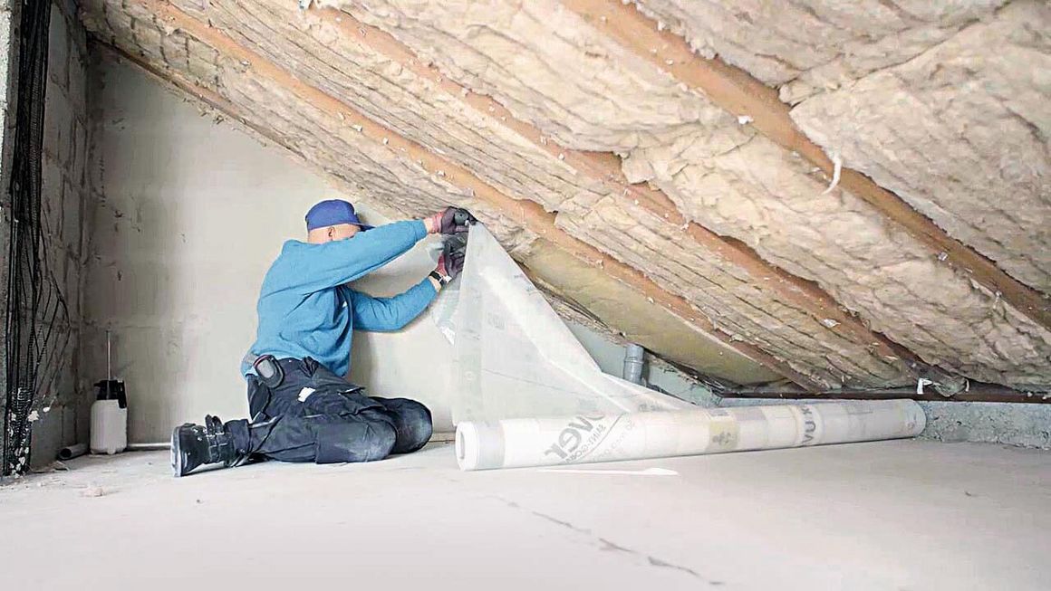 Ein Handwerker breitet eine Rolle Dampfbremsfolie auf dem Dachboden aus.