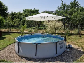 Ein runder Aufstellpool mit einem großen Sonnenschirm in einem sommerlichen Garten.