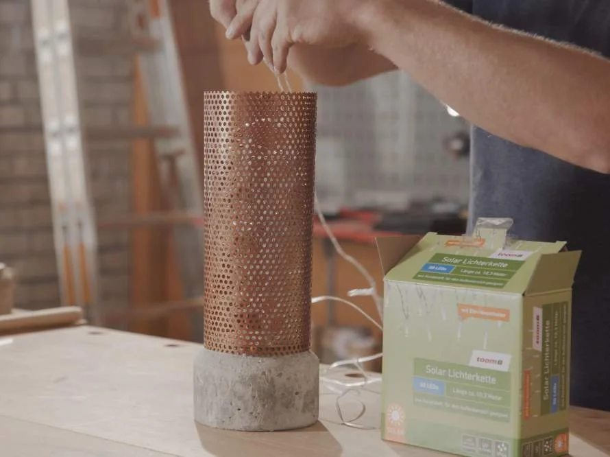 Person wickelt eine Lichterkette um eine Kupferröhre mit einem Betonfuß.