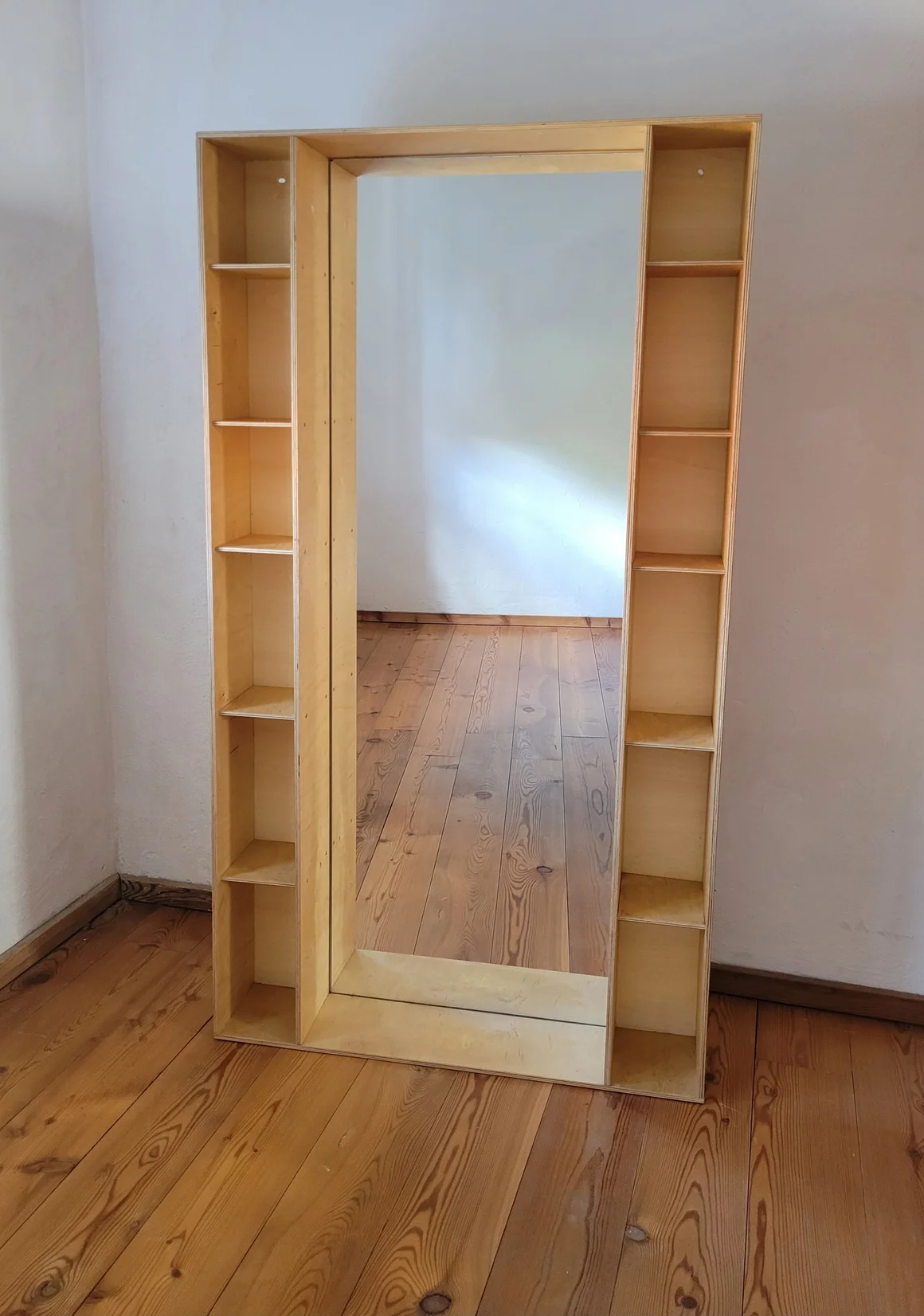 Ein Standspiegel mit seitlichen Regalen aus hellem Holz auf einem Holzboden.