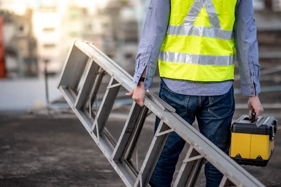 Person in Warnweste trägt Leiter und Werkzeugkasten.