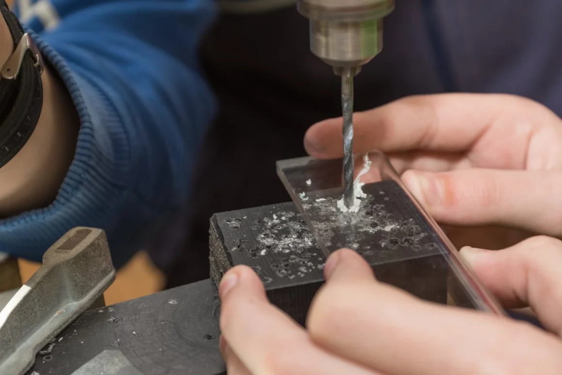 Eine Person bohrt mit einer Standbohrmaschine ein Loch in eine Acrylglasplatte.