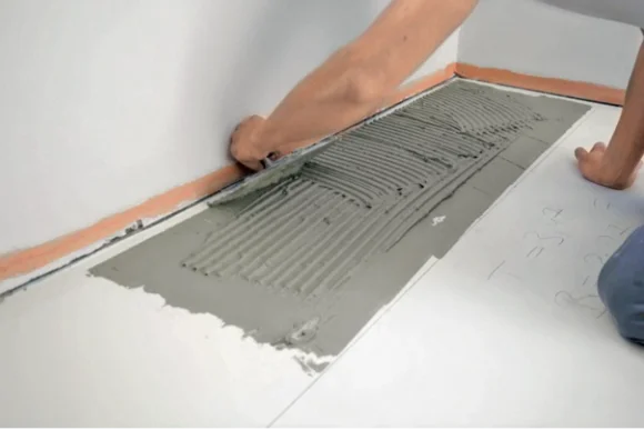 Eine Person trägt Fliesenkleber mit einer Zahnkelle auf dem Boden auf.