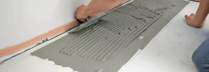 Eine Person trägt Fliesenkleber mit einer Zahnkelle auf dem Boden auf.