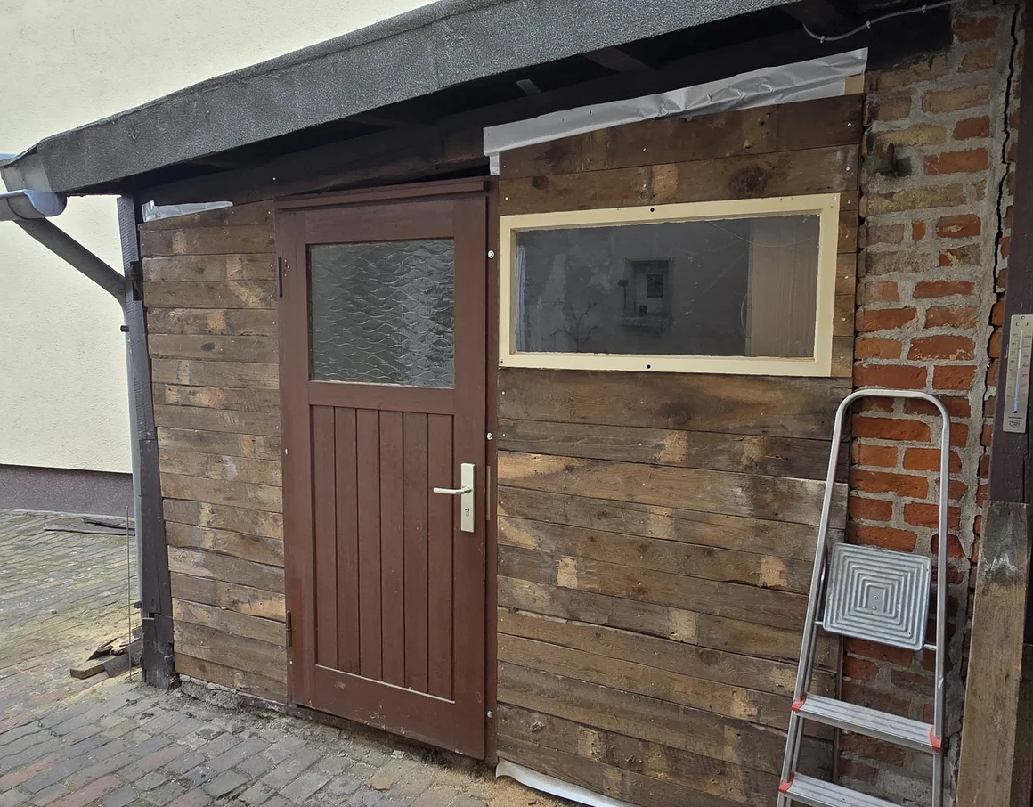 Ein unfertiger Holzschuppen mit Tür, Fenster und einer angelehnten Leiter.
