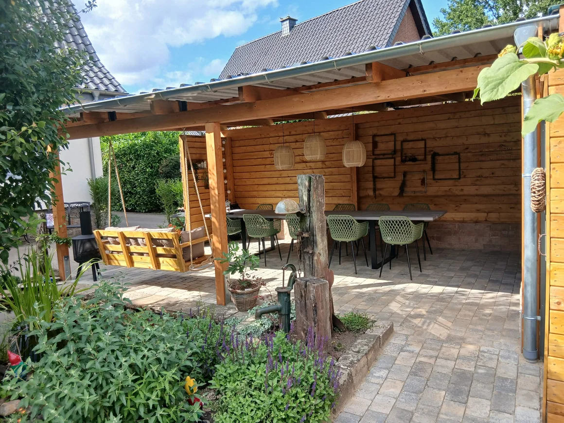Eine überdachte Holzterrasse mit großem Esstisch und einer Schaukelbank.