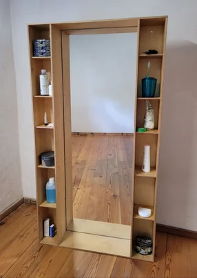 Ein Standspiegel aus Holz mit seitlichen Regalen voller Badutensilien.