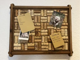 Eine Pinnwand aus Weinkorken, auf der ein Einkaufszettel, ein Notizzettel und zwei Fotos befestigt sind.