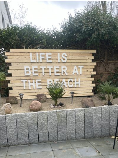 Holzschild mit der Aufschrift "Life is better at the beach" und zwei Palmen davor.
