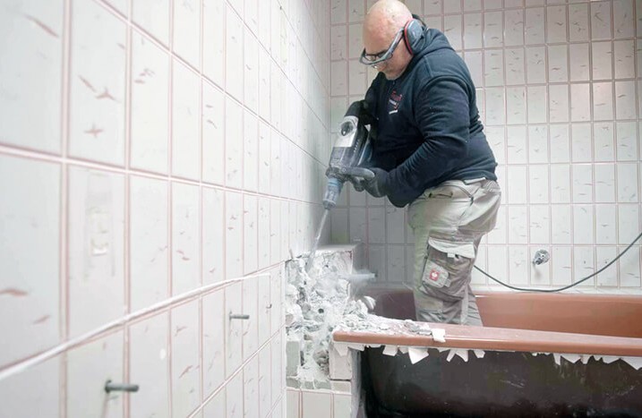 Ein Mann benutzt einen Stemmhammer zum Lösen der Fliesen um die Wanne.