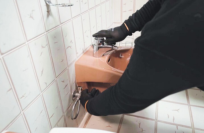 Ein Mann mit Handschuhen dreht Wasserzulauf am Waschbecken zu.