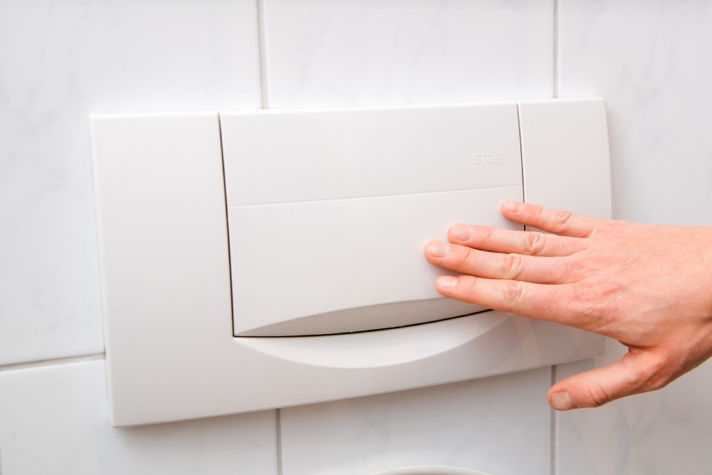 Eine Hand drückt die Spartaste einer Toilettenspülung an der Wand.