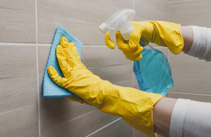 Eine Frau mit gelben Handschuhen reinigt eine Dusche mit einem Schwamm in der einen und Putzmittel in der anderen Hand.