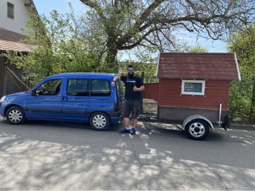 Person steht neben einem roten Mini-Holzhaus auf Rädern