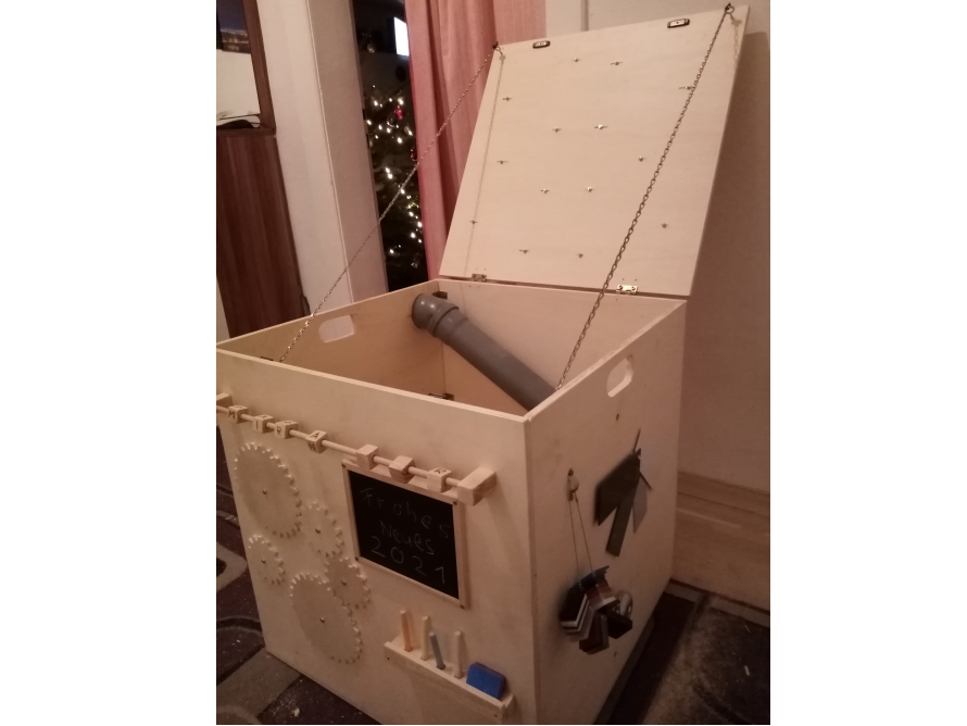 Eine Holzkiste mit Deckel, an der verschiedene motorische Spielelemente angebracht sind.