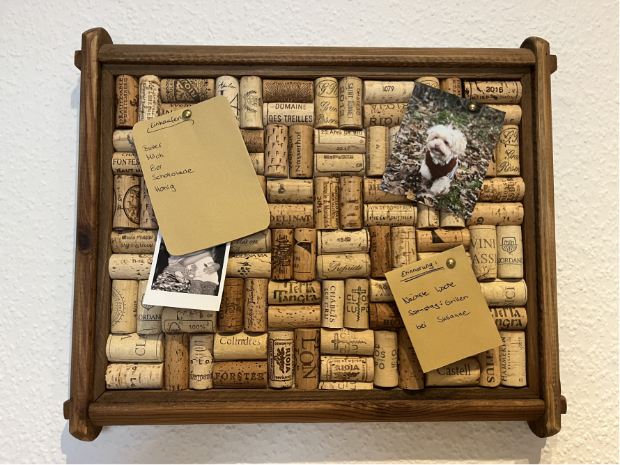 Eine Pinnwand aus Weinkorken, auf der ein Einkaufszettel, ein Notizzettel und zwei Fotos befestigt sind.