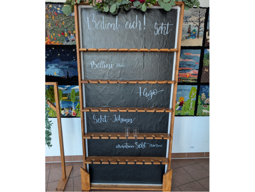 Ein hölzernes Regal mit mehreren Ebenen, in denen Sektgläser aufbewahrt werden können. Die Ebenen haben schwarze Tafeloberflächen, auf denen verschiedene Sektsorten aufgeschrieben sind.