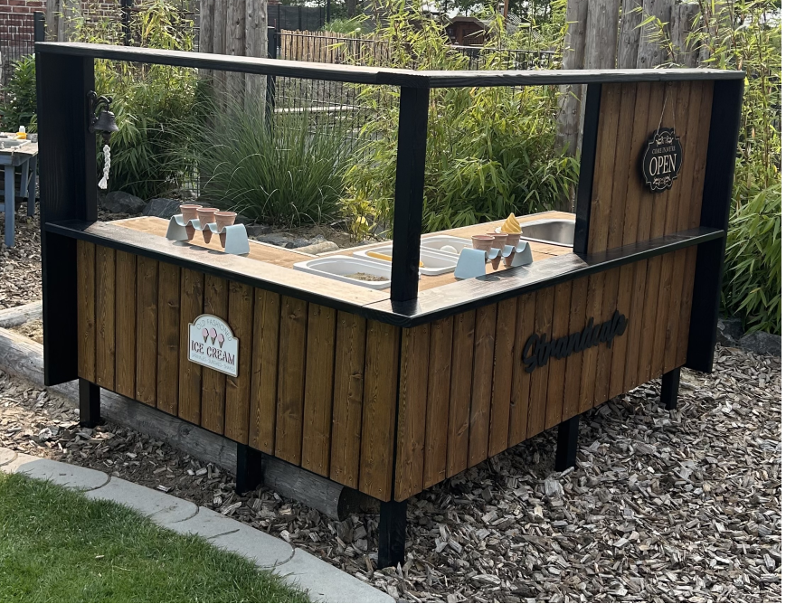 Eine Spiel-Eisdiele aus Holz steht in einem Garten auf Holzschnitzeln.