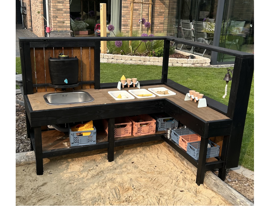 Eine schwarze, L-förmige Matschküche aus Holz steht in einem Sandkasten im Garten.