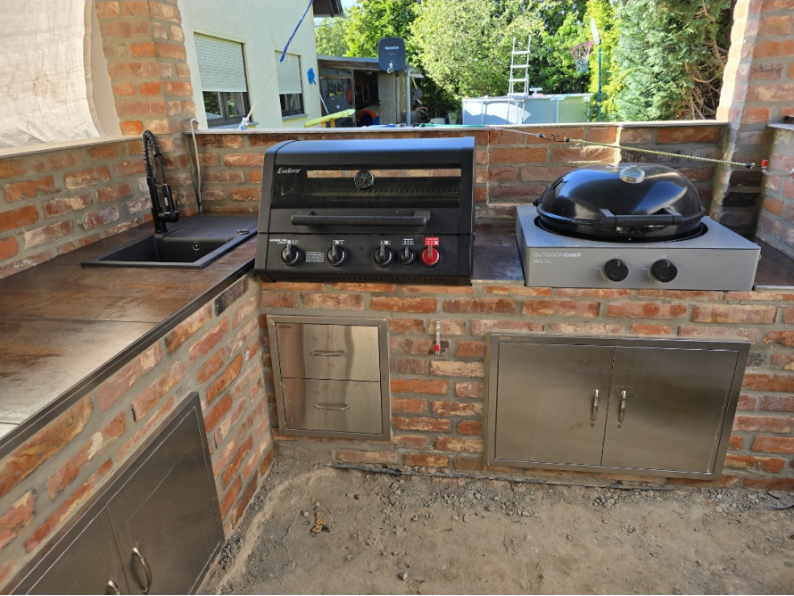 Eine gemauerte Außenküche mit eingebautem Grill, Spüle und Edelstahlschränken im Rohbau.