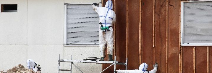 Personen in Ganzkörperschutzanzügen entfernen Asbest von einer Fassade.